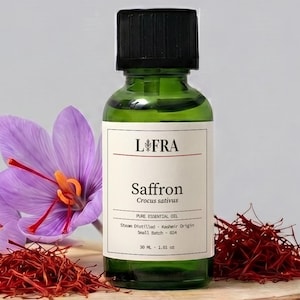 May include: A green glass bottle of L'FRA Saffron pure essential oil with a black cap, featuring a white label with black text. Beside the bottle are two purple crocus flowers and small piles of red saffron threads on a round wood slice.