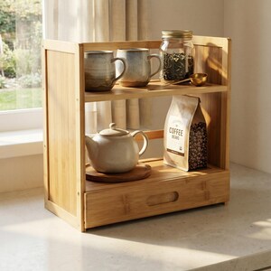 Bamboo counter organizer in sunlight with mugs and tea jar displayed on two shelves and drawer below.