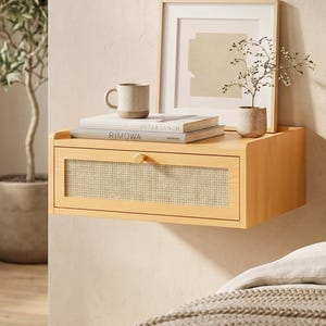 May include: A light wood floating bedside table with a woven cane drawer front and a brass knob. A framed print, a small potted plant, a mug, and books are on top. The table is mounted on a light-coloured wall.