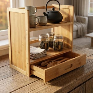 Bamboo two-tier organizer with drawer open on a wooden table holding tea jars and utensils.