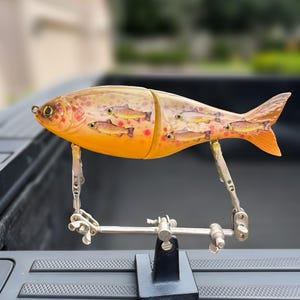 May include: A detailed, segmented fishing lure in the shape of a fish. The lure is primarily orange with brown and red spots, and features smaller fish designs inside. It is held by a silver metal stand.
