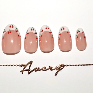 May include: Five oval false nails with a cherry design on a white tip. A gold-coloured chain necklace with the name "Avery" in cursive script is below the nails. The nails are in a row.