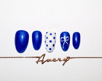 Pode incluir: Cinco unhas postiças azuis com acabamento metálico, uma com bolinhas brancas e outra com um desenho de laço branco. Uma corrente de ouro com o nome "Avery" está na parte inferior.