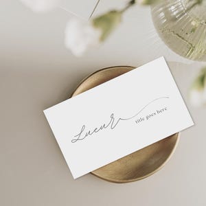 Puede incluir: Una tarjeta de visita blanca con la palabra "Lucur" en escritura cursiva y el texto "title goes here" descansa sobre un plato redondo dorado. El fondo es beige neutro con un arreglo floral borroso.