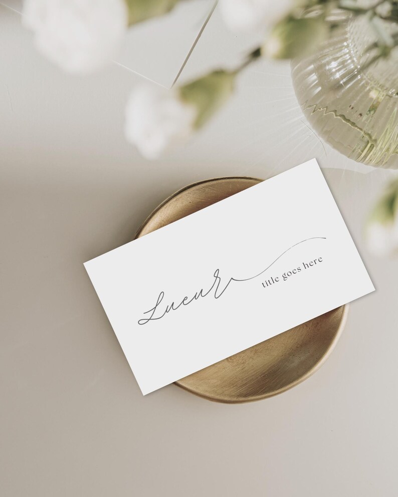 Puede incluir: Una tarjeta de visita blanca con la palabra "Lucur" en escritura cursiva y el texto "title goes here" descansa sobre un plato redondo dorado. El fondo es beige neutro con un arreglo floral borroso.