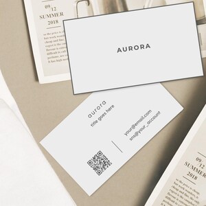 Puede incluir: Tarjetas de visita blancas con la palabra "AURORA" impresa en negro. Una tarjeta tiene un c&oacute;digo QR, correo electr&oacute;nico e informaci&oacute;n de redes sociales. Las tarjetas est&aacute;n sobre un fondo neutro con una p&aacute;gina de revista y una planta.