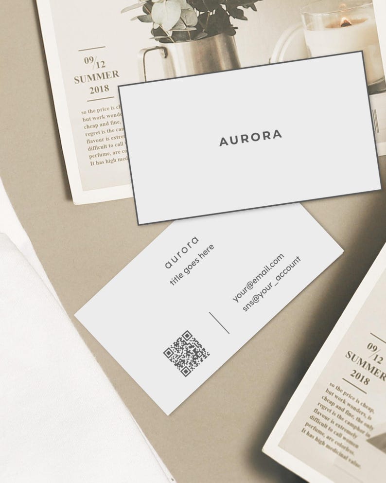 Puede incluir: Tarjetas de visita blancas con la palabra "AURORA" impresa en negro. Una tarjeta tiene un c&oacute;digo QR, correo electr&oacute;nico e informaci&oacute;n de redes sociales. Las tarjetas est&aacute;n sobre un fondo neutro con una p&aacute;gina de revista y una planta.