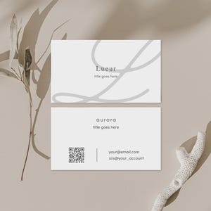 Puede incluir: Dos tarjetas de visita blancas con texto gris y un dise&ntilde;o decorativo gris. La tarjeta superior dice "Lueur" y "title goes here". La tarjeta inferior dice "aurora", "title goes here" e incluye un c&oacute;digo QR e informaci&oacute;n de contacto.