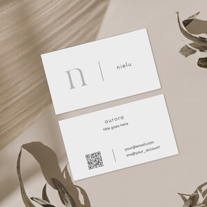 Puede incluir: Dos tarjetas de visita blancas con dise&ntilde;o minimalista. La tarjeta superior presenta el nombre "nielu" y una letra "n" estilizada. La tarjeta inferior incluye la palabra "aurora", el texto "title goes here", un c&oacute;digo QR e informaci&oacute;n de contacto.