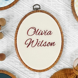 Puede incluir: Un bastidor de bordado ovalado con acabado de imitación madera que contiene una tela de color crema. El nombre Olivia Wilson está bordado en el centro con hilo rojo oscuro en letra cursiva. Incluye un gancho metálico para colgarlo.