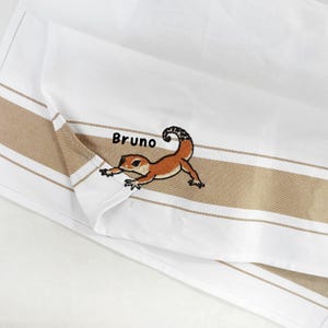 Peut inclure: Un torchon de cuisine blanc avec une bande horizontale beige et une broderie représentant un lézard orange à la queue recourbée. Le prénom Bruno est brodé en fil noir au-dessus du lézard. Le tissu présente une texture tissée et des bords ourlés.