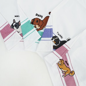 May include: Four white kitchen towels featuring embroidered dog illustrations and names. Each towel has a coloured striped border in red, teal, blue, or pink. The dogs are labelled Max, Daisy, Teddy, and Charlie, with detailed stitching for each breed.