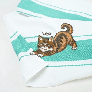 Pode incluir: Um pano de prato branco com listras horizontais verde-azuladas e um bordado de um gato malhado marrom e branco em posição de alongamento. O nome Leo está bordado em letras pequenas marrons acima do gato, que tem patas brancas.