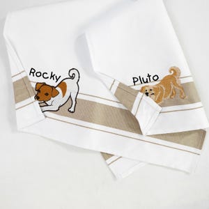 May include: A white kitchen towel featuring a horizontal tan stripe and embroidered dog designs. One side shows a brown and white dog with the name Rocky embroidered above it, while the other side shows a golden dog with the name Pluto embroidered above it.