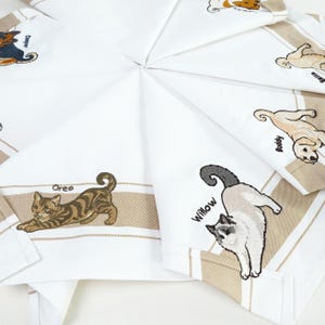 Könnte beinhalten: Ein Set aus weißen Stoffservietten mit gestickten Illustrationen verschiedener Katzen und Hunde. Jedes Tier wird von einem in schwarzem Garn gestickten Namen begleitet, darunter Oreo, Willow, Cooper und Buddy, auf einem beigen Streifenrand.