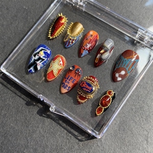 May include: A collection of custom-designed nail art in a clear plastic case. The nails feature various designs, including portraits, geometric patterns, and the text "Life on Mars" and "Bowie". Colors include red, blue, gold, and brown.