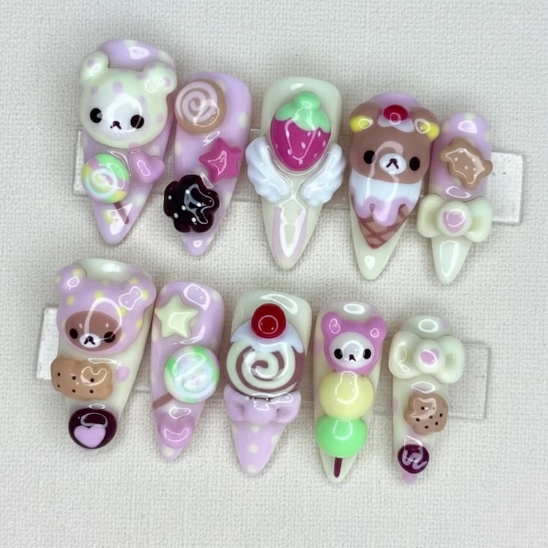 May include: A set of ten artificial nails with a light pink base, each adorned with 3D designs. The designs include teddy bears, lollipops, strawberries with wings, ice cream cones, and bows. The nails are decorated with various colors.