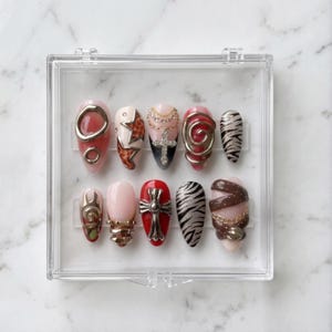 May include: A set of ten artificial nails in a clear plastic case. The nails feature various designs, including gold accents, animal prints, and cross motifs. Colors include red, pink, black, and brown. The nails are oval-shaped and appear to be professionally designed.