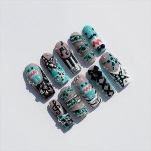 May include: A collection of long, decorated artificial nails. Designs include musical notes, bows, and cartoon characters in teal, black, and silver. Some nails feature rhinestones and intricate patterns, set against a white background.