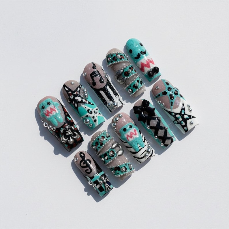 May include: A collection of long, decorated artificial nails. Designs include musical notes, bows, and cartoon characters in teal, black, and silver. Some nails feature rhinestones and intricate patterns, set against a white background.