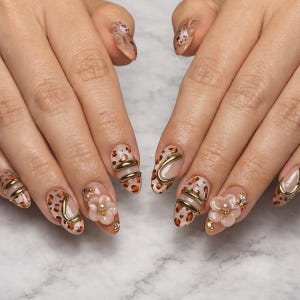 Puede incluir: Uñas manicuradas con una base nude y patrones de estampado de leopardo marrón, acentuadas con barras metálicas doradas curvas, pequeños adornos florales blancos con perlas centrales y diminutas cuentas doradas en la superficie.