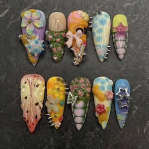 May include: A set of ten press-on nails featuring diverse 3D designs including pastel flowers, pink bows, silver star charms, green moss-like textures, blue water-like patterns, and colorful resin accents on almond and stiletto shaped nail tips.