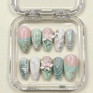 Puede incluir: Un juego de diez uñas artificiales en una caja de plástico transparente. Las uñas presentan varios diseños, incluyendo peces koi, patrones florales y texto japonés. Los colores incluyen rosa, verde, blanco e iridiscente. Las uñas son de forma ovalada.