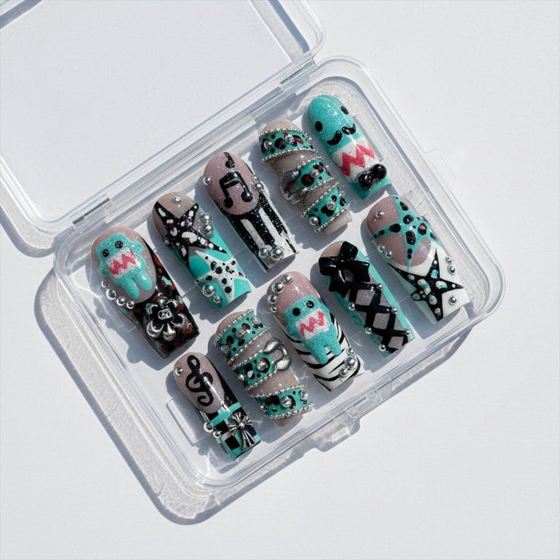 May include: A set of ten artificial nails in a clear plastic case. The nails feature various designs, including musical notes, abstract patterns, and cartoon characters, with accents of teal, black, and silver. Each nail is uniquely decorated.