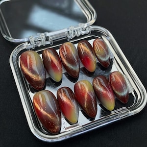 May include: A set of ten oval-shaped false nails in a clear plastic case. The nails feature a gradient design with colors ranging from deep red to gold and silver, creating a cat-eye effect. The nails are glossy and appear ready for application.