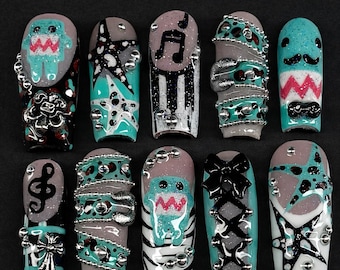 Op de afbeelding: Een verzameling van tien kunstnagels met diverse ontwerpen. De nagels tonen tinten van teal, grijs, zwart en zilver, met versieringen zoals muzieknoten, sterren en cartoonfiguren. Sommige nagels hebben een getextureerd uiterlijk, andere ingewikkelde patronen.