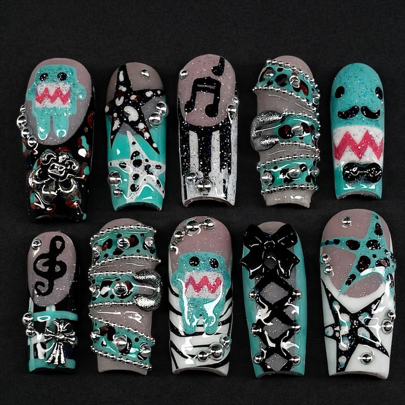 May include: A collection of ten artificial nails with various designs. The nails feature a range of colors, including teal, gray, black, and silver, with embellishments like musical notes, stars, and cartoon characters. Some nails have a textured appearance, while others showcase intricate patterns.
