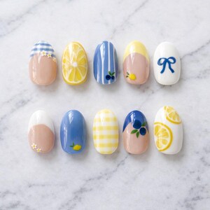 Puede incluir: Un conjunto de diez uñas postizas ovaladas con varios diseños que incluyen cuadros vichy azules y blancos, rodajas de limón amarillo, rayas verticales azules con arándanos, puntas amarillas con limones, lazos azules, flores y azul sólido.