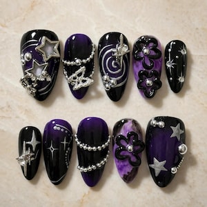 May include: A set of ten almond-shaped press-on nails featuring a black and purple color palette. The designs include silver star charms, metallic bead chains, raised black flower motifs, swirling patterns, and small rhinestones on a glossy finish.