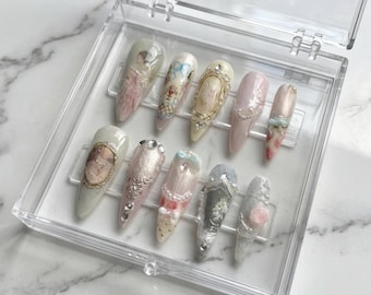 May include: A set of ten almond-shaped press-on nails displayed in a clear plastic case. Each nail features intricate 3D embellishments, including gold frames, miniature pearls, floral patterns, and cameo-style portraits in soft pastel tones.