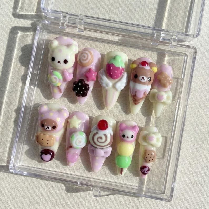 May include: A set of ten artificial nails in a clear plastic case. Each nail is decorated with a different design, including bears, strawberries, and sweets. The nails are primarily pink, white, and brown, with various other colors used for accents.