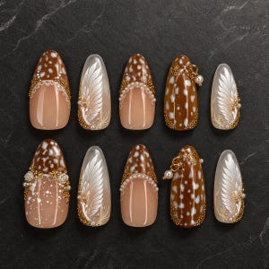 Op de afbeelding: Een set van tien amandelvormige kunstnagels met een mix van bruine en witte vlekpatronen, witte veerachtige ontwerpen, kleine witte bloemaccenten, gouden metalen details en kleine parelkralen, waarbij sommige nagels kleine gouden ringen bevatten.