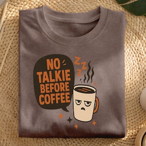 May include: A brown t-shirt with the slogan "NO TALKIE BEFORE COFFEE" in an orange speech bubble. A cartoon coffee cup with a grumpy face and steam rising from it is also featured. The shirt is folded on a woven mat.
