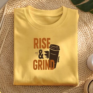 May include: Mustard yellow t-shirt featuring the text "RISE & GRIND" and a brown coffee cup graphic. The shirt is folded on a woven mat with decorative items and a monstera leaf.