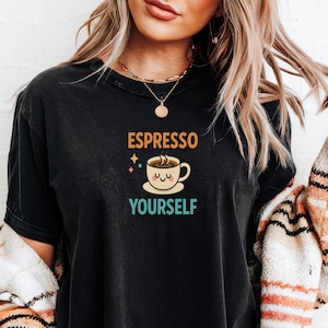 May include: Black t-shirt featuring the phrase "ESPRESSO YOURSELF" above a cartoon coffee cup. The coffee cup is light brown with a smiling face and steam rising from it. The text is in orange and teal.