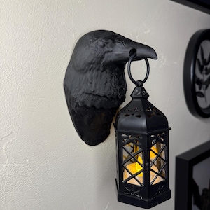 Könnte beinhalten: Eine schwarze Rabenkopf-Wandleuchte mit einer hängenden schwarzen Laterne. Die Laterne hat ein dekoratives Gitterdesign und enthält eine flackernde, warm getönte LED-Kerze. Der Rabenkopf ist an einer strukturierten weißen Wand befestigt.