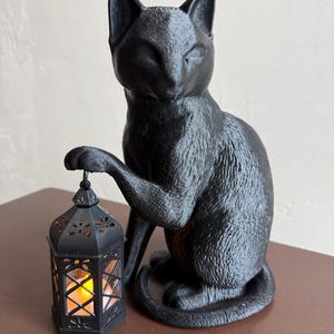 Pode incluir: Uma estatueta de gato preto segurando uma lanterna decorativa com uma luz tremeluzente semelhante a uma vela. O gato está sentado com a pata apoiada na lanterna. A lanterna tem um design geométrico.