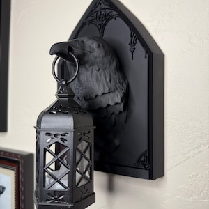 Könnte beinhalten: Schwarze Raben-Wandleuchte mit gotischem Bogenrücken. Der Rabe hält einen Metallring, der eine schwarze Laterne mit Glasscheiben trägt. Die Laterne hat eine Kerze im Inneren, was eine gruselige, gotische Ästhetik erzeugt.