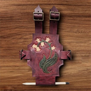 May include: Burgundy leather journal cover with a floral design, featuring a lily of the valley. The cover has adjustable straps with brass buckles and a pencil holder. The journal cover is on a wooden surface.