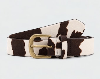 May include: A belt featuring a brown and white cow print pattern with a textured surface. The belt includes a single prong buckle made of brushed brass metal, a matching belt loop, and a series of pre-punched holes for adjustable fastening.