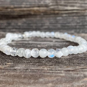 May include: A white moonstone bracelet with a silver clasp. The bracelet is made of round beads and is lying on a wooden surface.