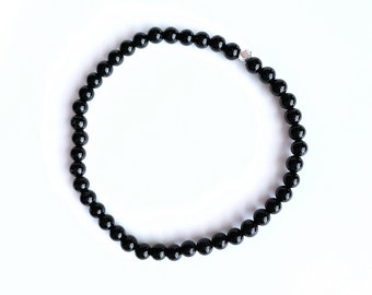 May include: A black beaded bracelet with small, round beads. The bracelet is strung together and features a small silver-colored accent bead. The beads are glossy and reflect light, creating a polished look. The bracelet is displayed on a white background.