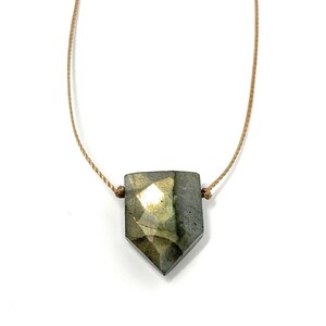 May include: A faceted, shield-shaped pendant necklace. The pendant is a mix of gray, green, and gold tones, suspended from a thin, tan cord. The necklace is displayed against a white background, highlighting the gemstone's unique shape and color.