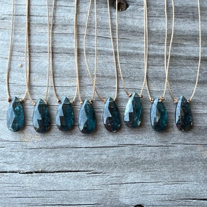 May include: Ten teal blue gemstone pendants on brown string necklaces. The gemstones are faceted and have a teardrop shape.