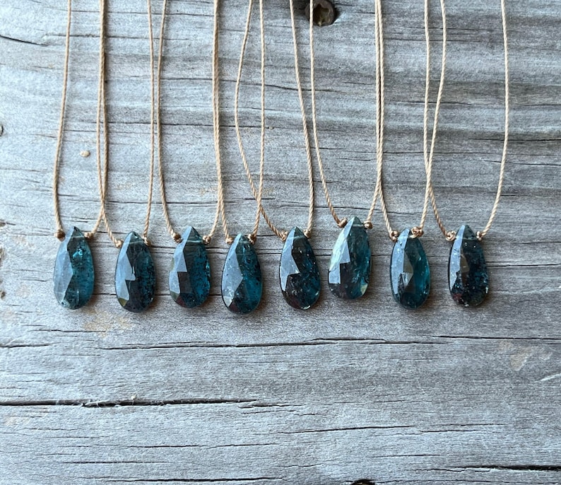 May include: Ten teal blue gemstone pendants on brown string necklaces. The gemstones are faceted and have a teardrop shape.