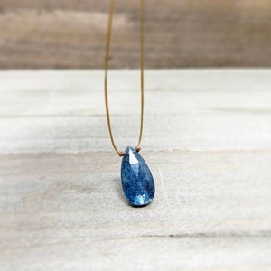May include: A teardrop-shaped blue gemstone pendant on a thin brown cord necklace.